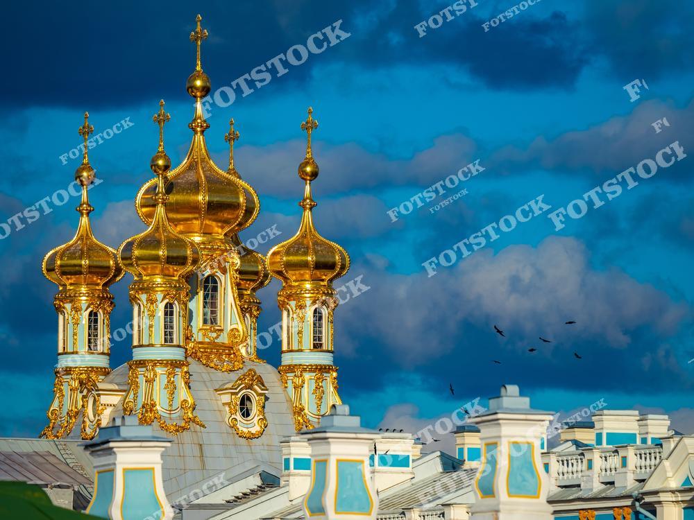 На фотографии изображены золотые купола дворцовой церкви Екатерининского дворца в Царском Селе. Эти купола, выполненные в стиле русского барокко, являются одной из ярчайших деталей комплекса и символом императорской роскоши. Темные облака на фоне подчеркивают блеск золота и добавляют драматичности изображению. Екатерининский дворец - выдающийся памятник архитектуры, привлекающий множество туристов своей историей и красотой.